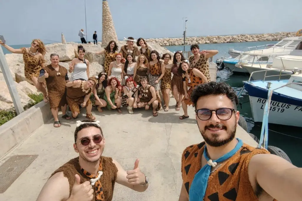 Grup mare de persoane pozând pentru un selfie în aer liber, pe un ponton lângă mare. Participanții poartă costume tematice inspirate din epoca preistorică (tip „Flintstones”), cu imprimeuri animale și accesorii din „oase”. În prim-plan, două persoane zâmbesc și fac semn cu degetul mare în sus. În fundal se văd bărci, apă și o zonă de dig din piatră, într-o zi însorită.
