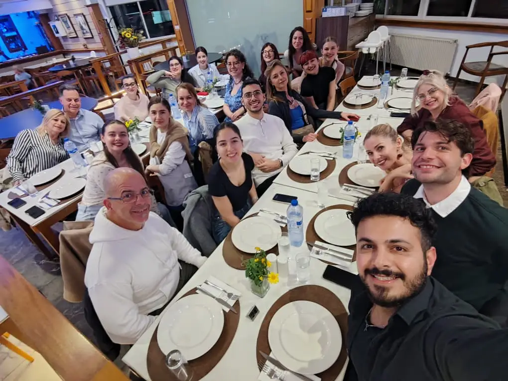 Grup de aproximativ 15–20 de persoane așezate la o masă lungă într-un restaurant, pozând pentru o fotografie de tip selfie. Toți zâmbesc către cameră, într-o atmosferă relaxată. Masa este pregătită cu farfurii, tacâmuri, pahare și sticle de apă, iar în mijloc sunt mici aranjamente florale. Interiorul este luminos, cu mobilier din lemn și decor simplu.
